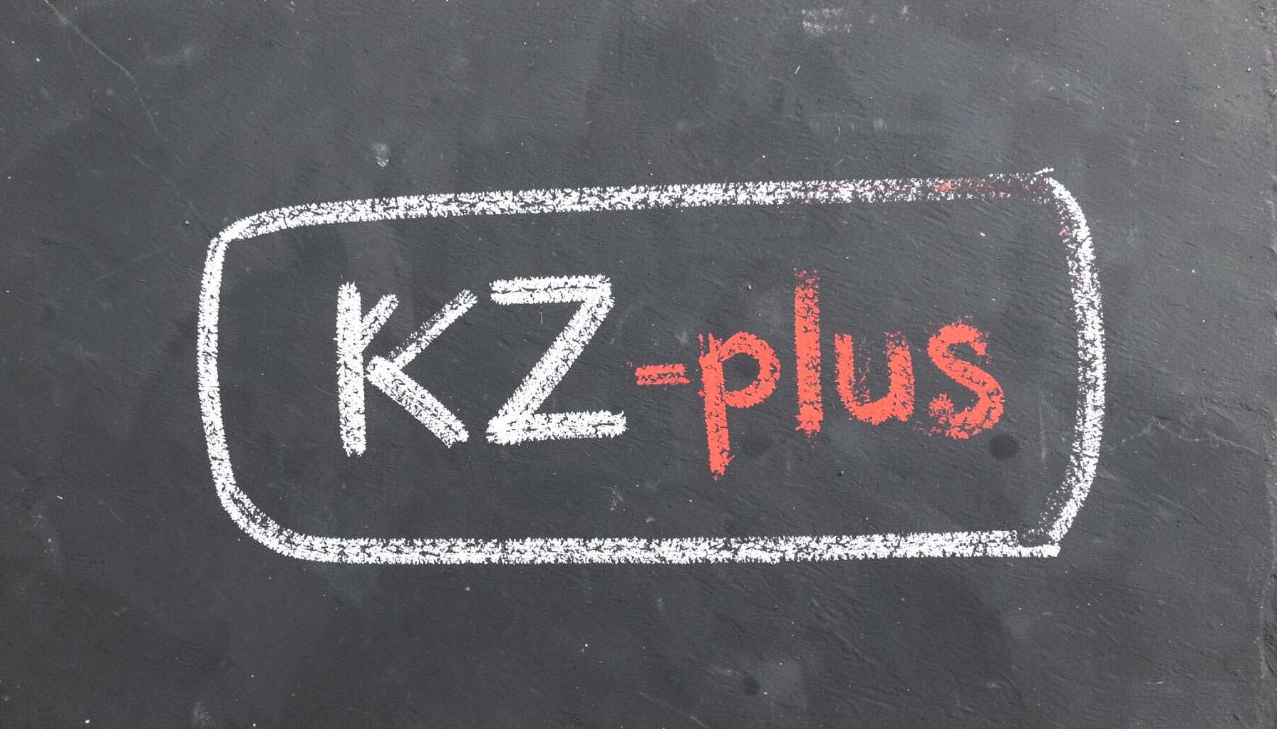 Меловой рисунок на тёмной грифельной доске: контур play-кнопки и надпись KZ-plus, выполненные белым и красным мелом на сером фоне.